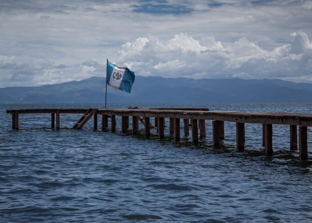 Morar na Guatemala em 2026: Custos, Visto e Realidade Areal e cais de madeira no Lago Atitlán com a bandeira da Guatemala a hastear sob céu nublado.