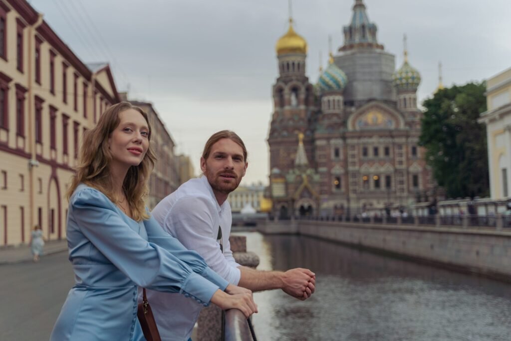 Casal jovem sorrindo ao lado de um canal em São Petersburgo com a Igreja do Sangue Derramado ao fundo.
