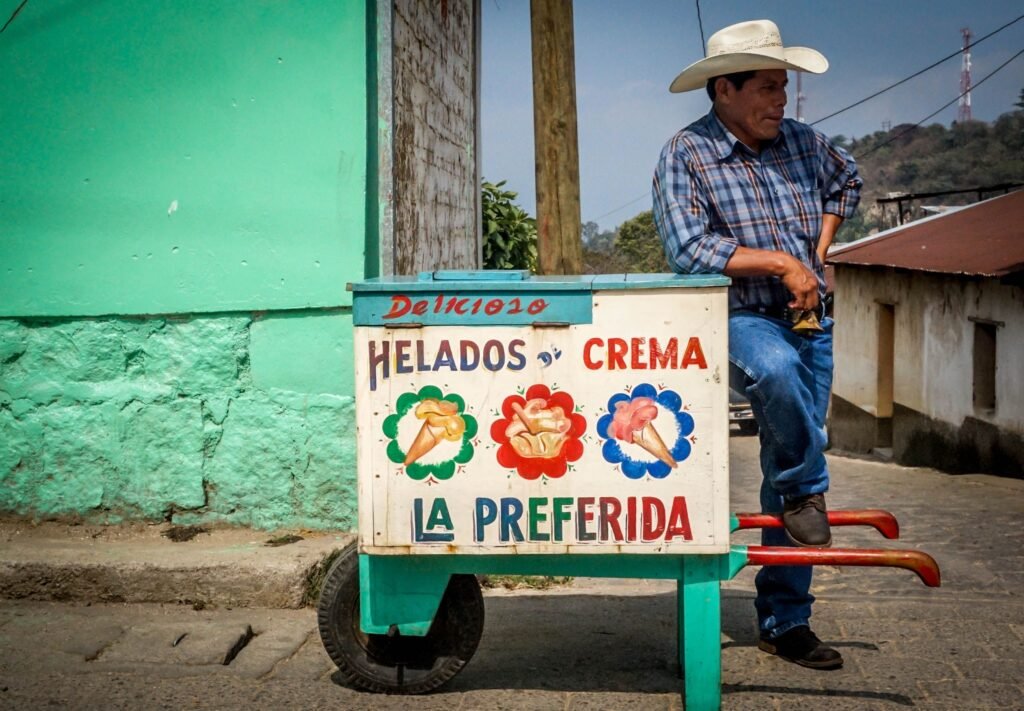 Trabalhar na Guatemala em 2026: Vistos, Salários e Vagas Homem de chapéu encostado em muro colorido ao lado de uma banca tradicional de gelados.