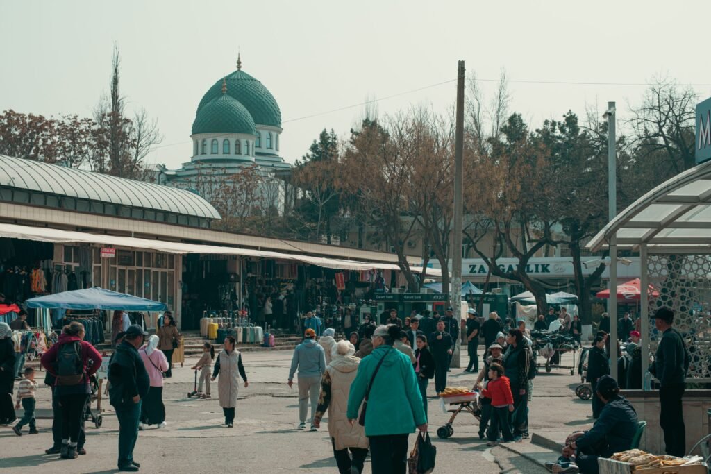 Morar no Uzbequistão em 2026: Custos, Visto e Realidade Área comercial e mercado local com vegetação, mesquita de cúpula verde ao fundo e pessoas a circular no Uzbequistão.